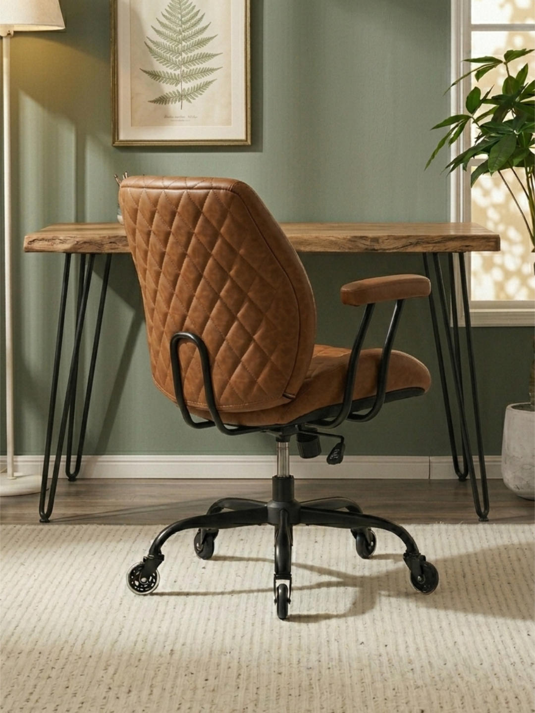 A warm brown leather desk chair with a quilted backrest, black metal base, and quiet casters stands in front of a live-edge wood desk with hairpin legs against a sage green wall.