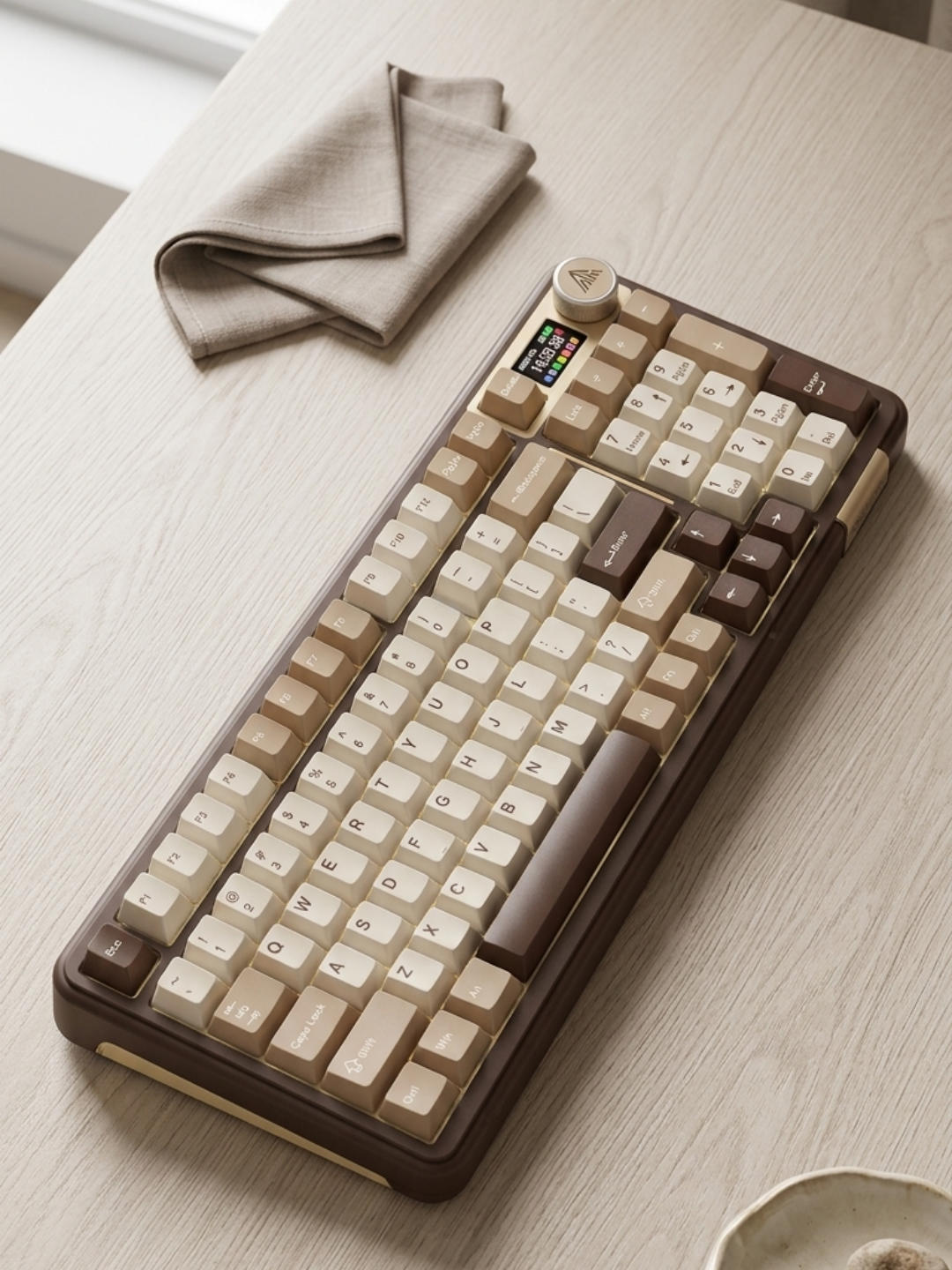 A wireless mechanical keyboard with cream, tan, and dark brown keys sits on a light wood desk next to a folded beige linen cloth.