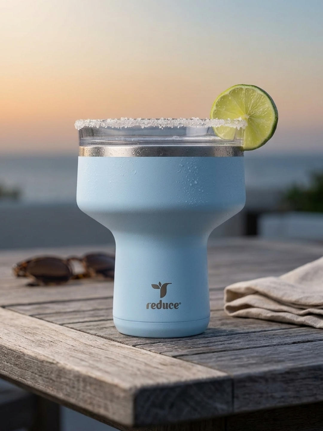A light blue insulated cocktail tumbler shaped like a stemless martini glass sits on a weathered wooden table. The rim is rimmed with salt and decorated with a lime wheel. Sunglasses and a linen napkin rest nearby under a warm sunset sky.