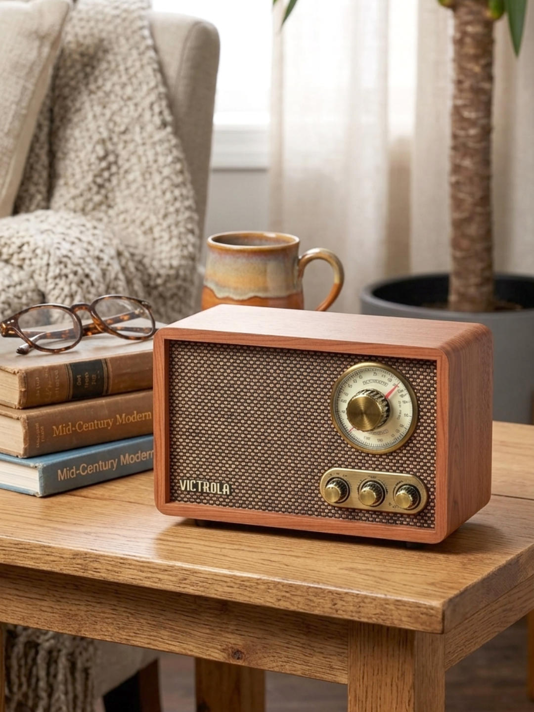 A rectangular wood-cased radio sits on a light oak tabletop next to stacked books, a pair of glasses, and a ceramic mug. The radio has a walnut finish, a woven brown speaker grill, a large circular gold-colored dial, and three smaller gold knobs.