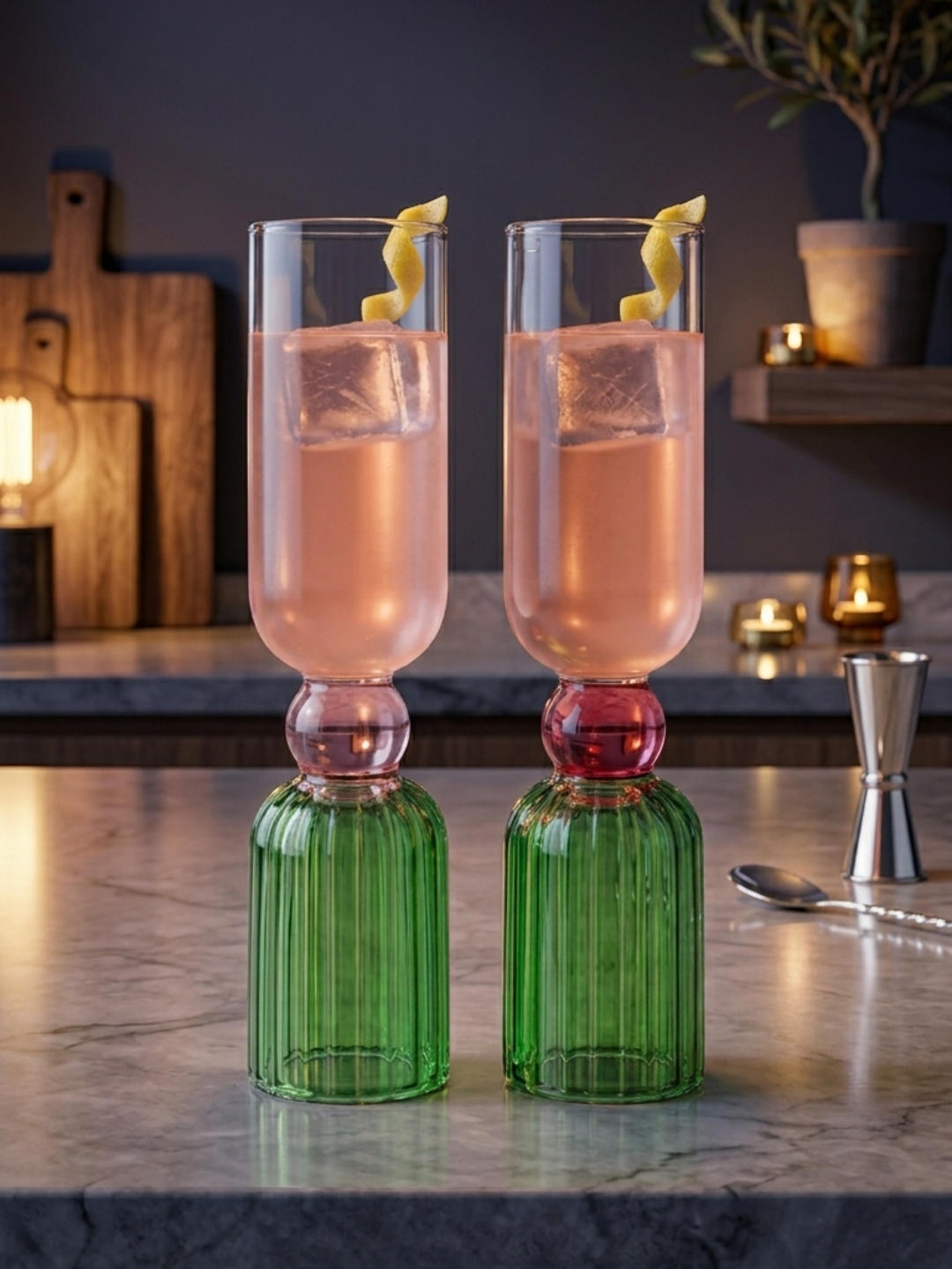 Two tall, reversible cocktail glasses filled with a pink beverage and a lemon twist garnish, standing on a marble countertop. The bottom portion is ridged green glass, the middle is a smooth pink glass sphere, and the top is clear glass.