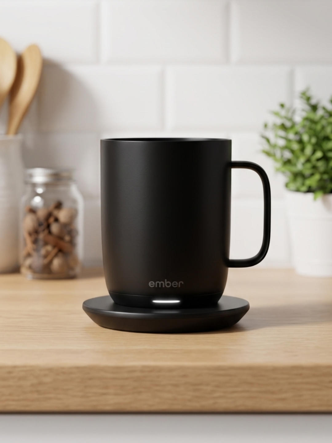A matte black Ember smart mug sits on its matching black charging saucer on a light wooden countertop. In the background, a white tiled wall, a glass jar of spices, and a small potted plant are softly blurred.