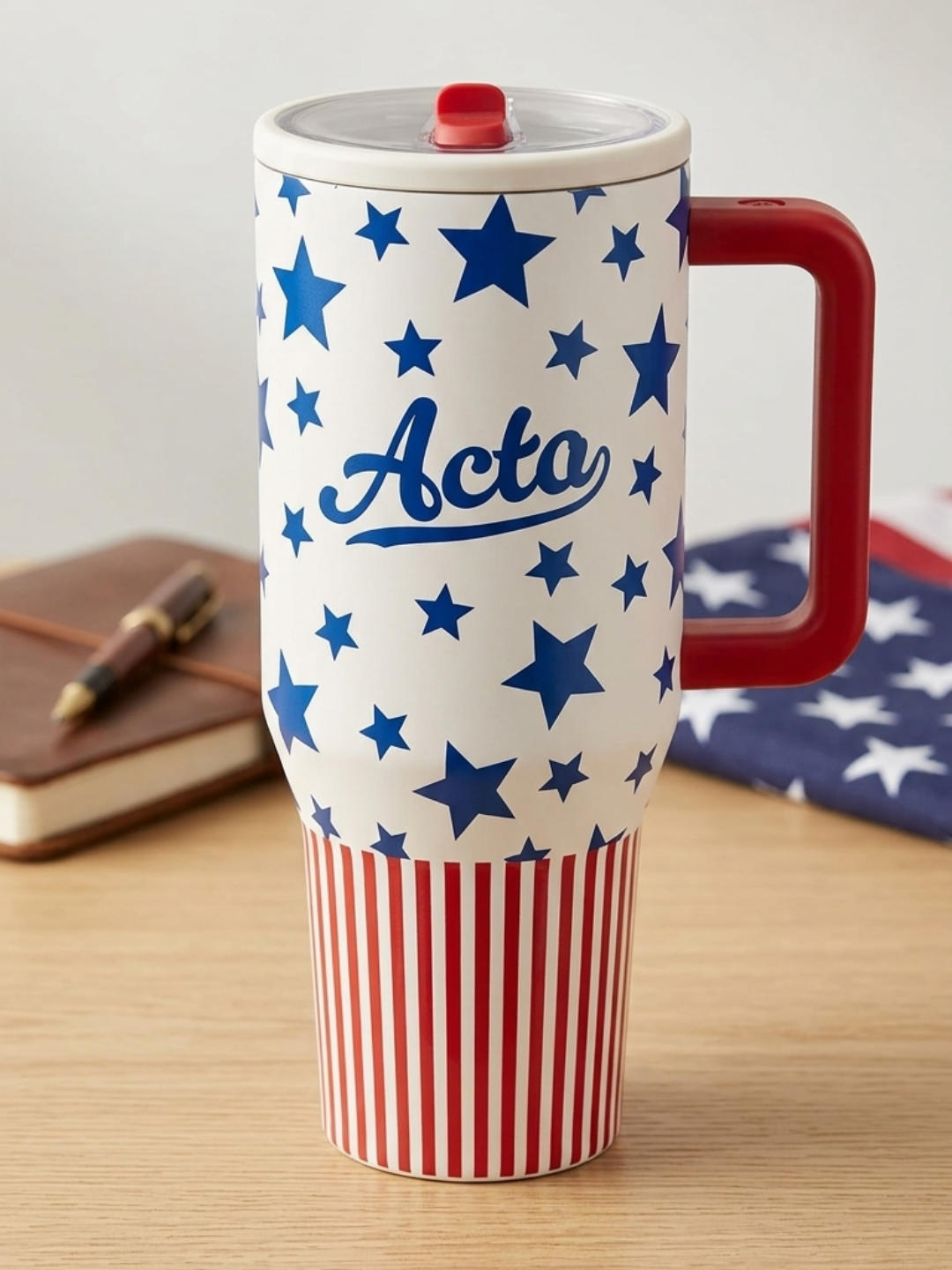 A tall insulated tumbler with a white upper body covered in blue stars and a blue script logo, a tapered lower body with red and white vertical stripes, a solid red handle, and a clear lid with a red flip tab, sitting on a light wooden table next to a brown leather notebook and a folded American fla