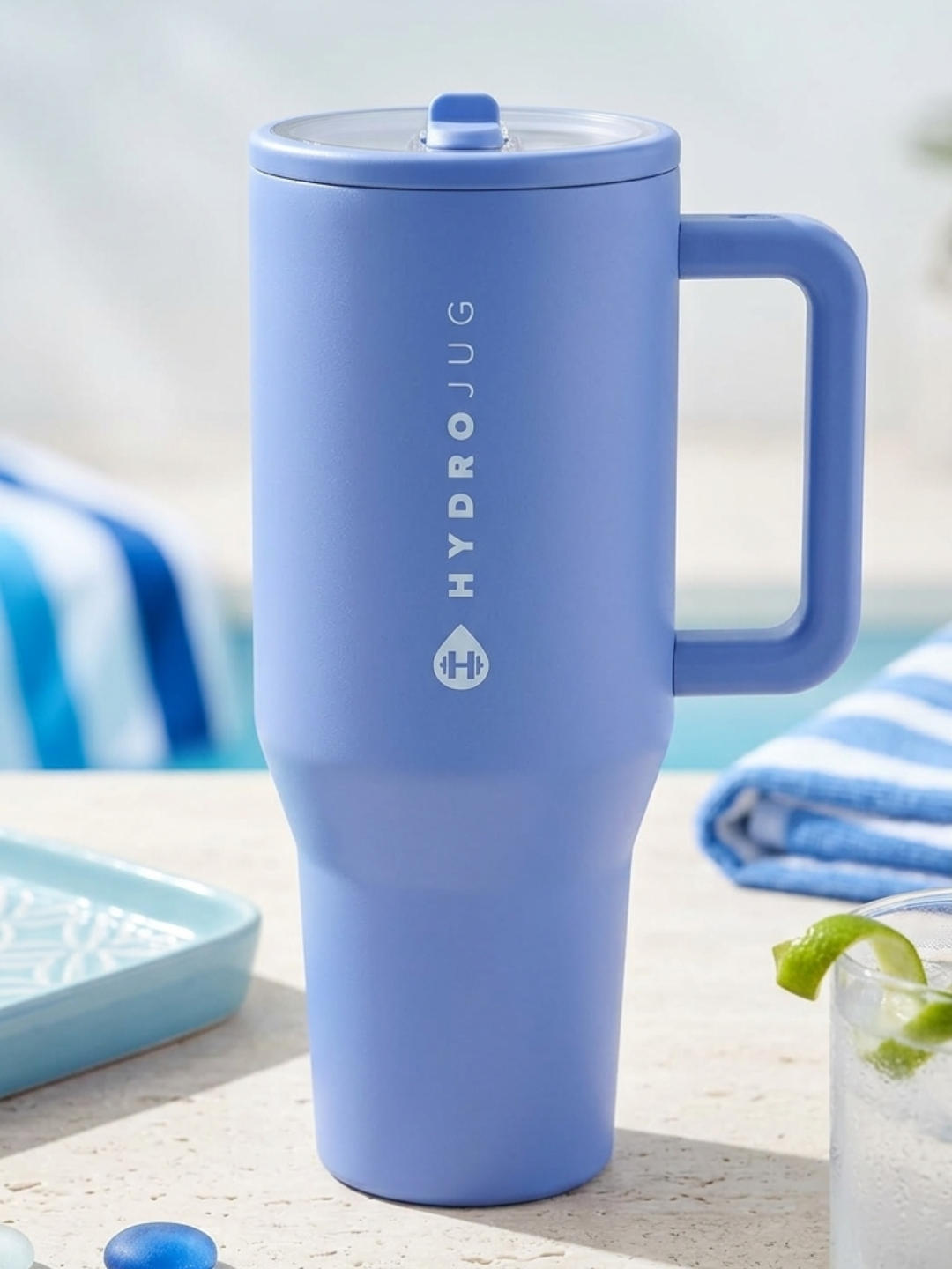 A light blue insulated travel tumbler with a sturdy square handle sits on a light stone surface, with a glass of water and blue striped towels visible in the background.