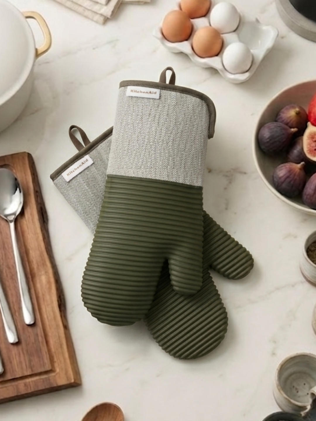 A pair of forest green and grey herringbone oven mitts lying crossed on a light marble countertop next to a wooden cutting board, silver spoons, a bowl of fresh figs, and a small carton of brown and white eggs.