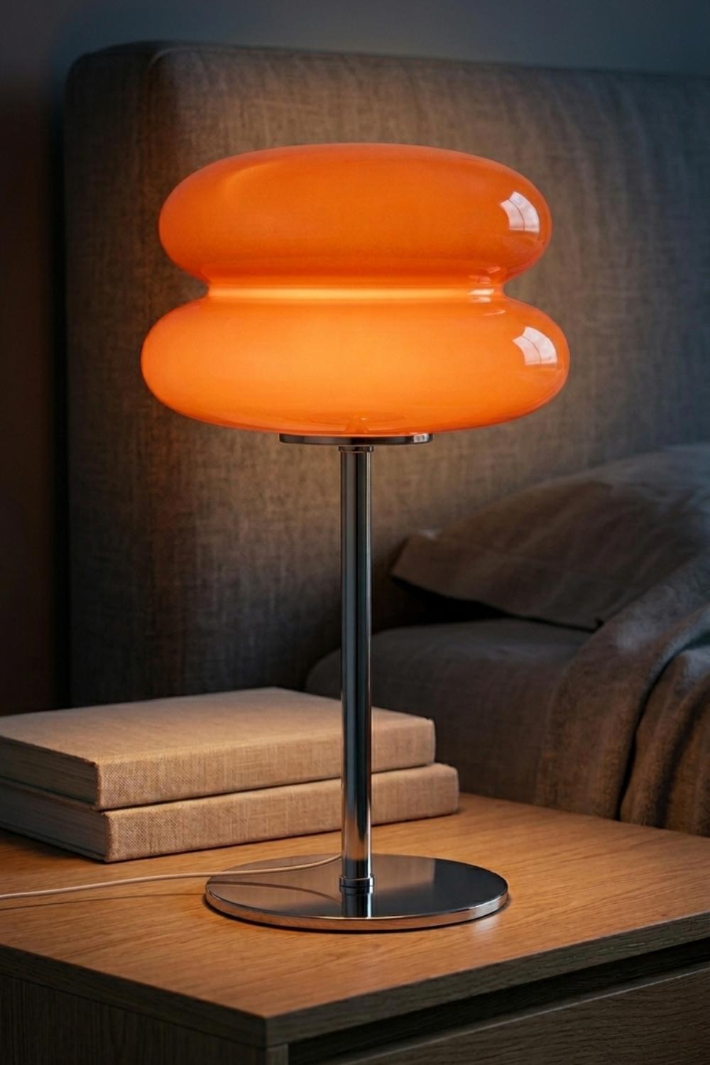 An illuminated orange glass table lamp with a mushroom-shaped shade and a polished silver metal base, sitting on a wooden nightstand next to two stacked books in front of a neutral upholstered bed headboard.