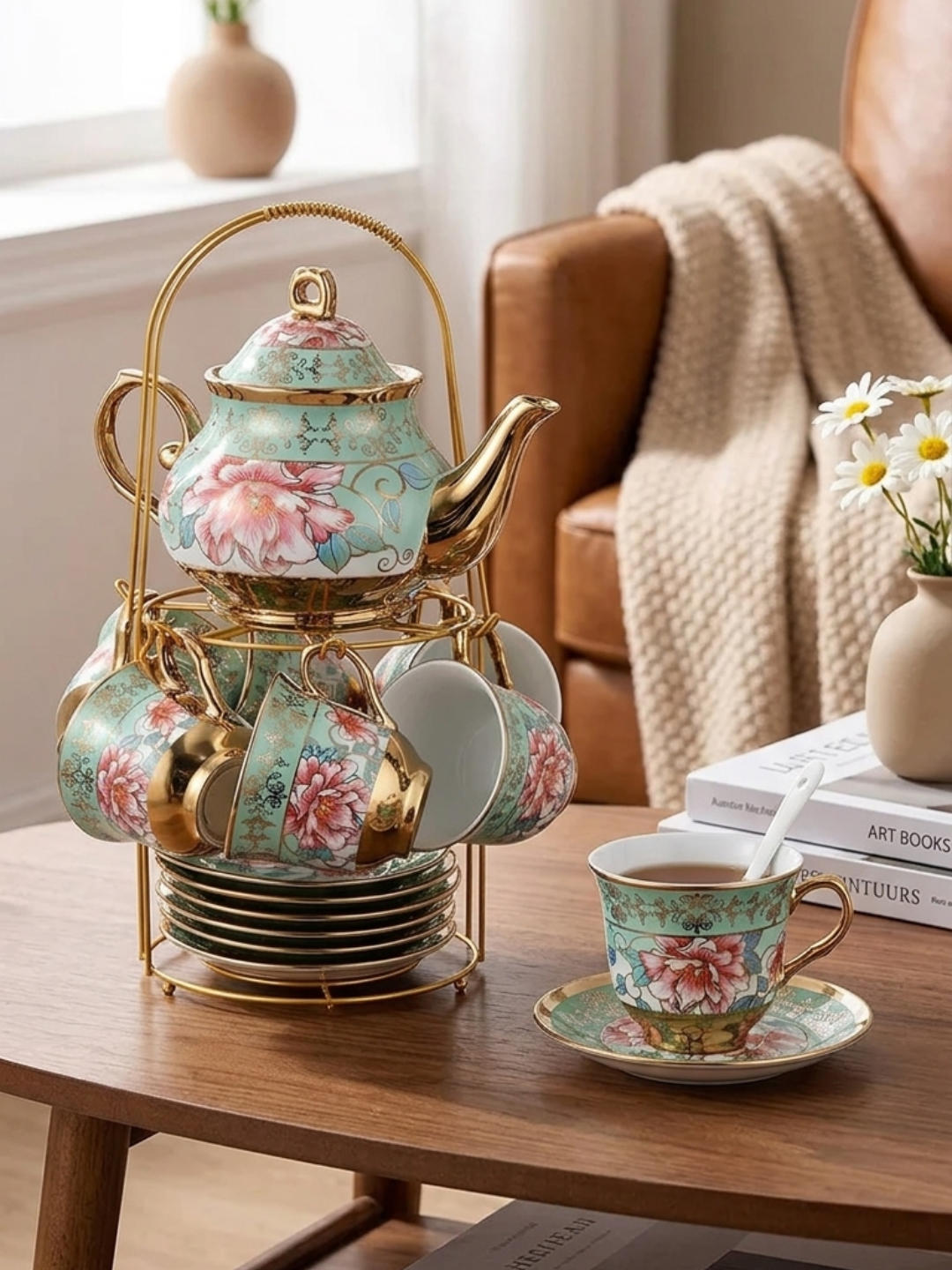 A mint green porcelain tea set with pink floral patterns and gold trim, displayed on a gold-toned wire stand on a wooden coffee table. One cup with a spoon is placed on its saucer next to the stand, with books and a vase of daisies in the background.