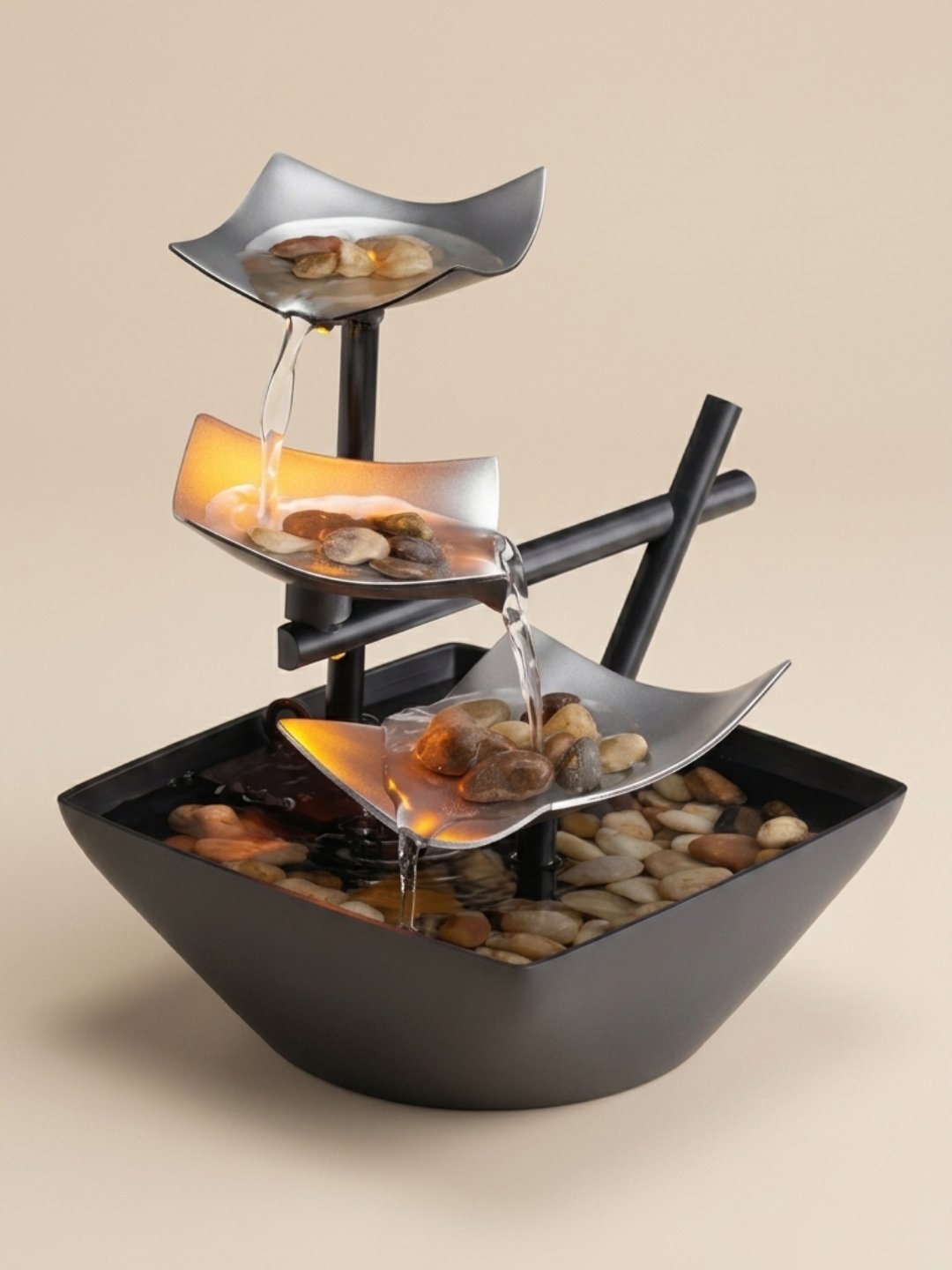 A three-tier indoor tabletop water fountain with curved silver leaves pouring water over smooth river rocks into a dark square basin, softly illuminated by warm amber light.