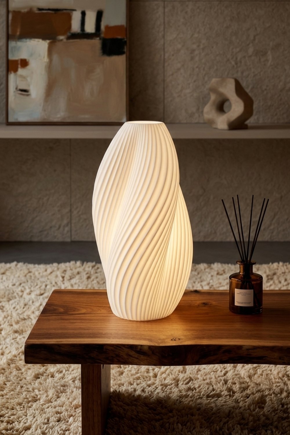 A lit sculptural table lamp with a twisted fluted white body casting a warm glow on a live-edge wooden table, beside an amber reed diffuser.