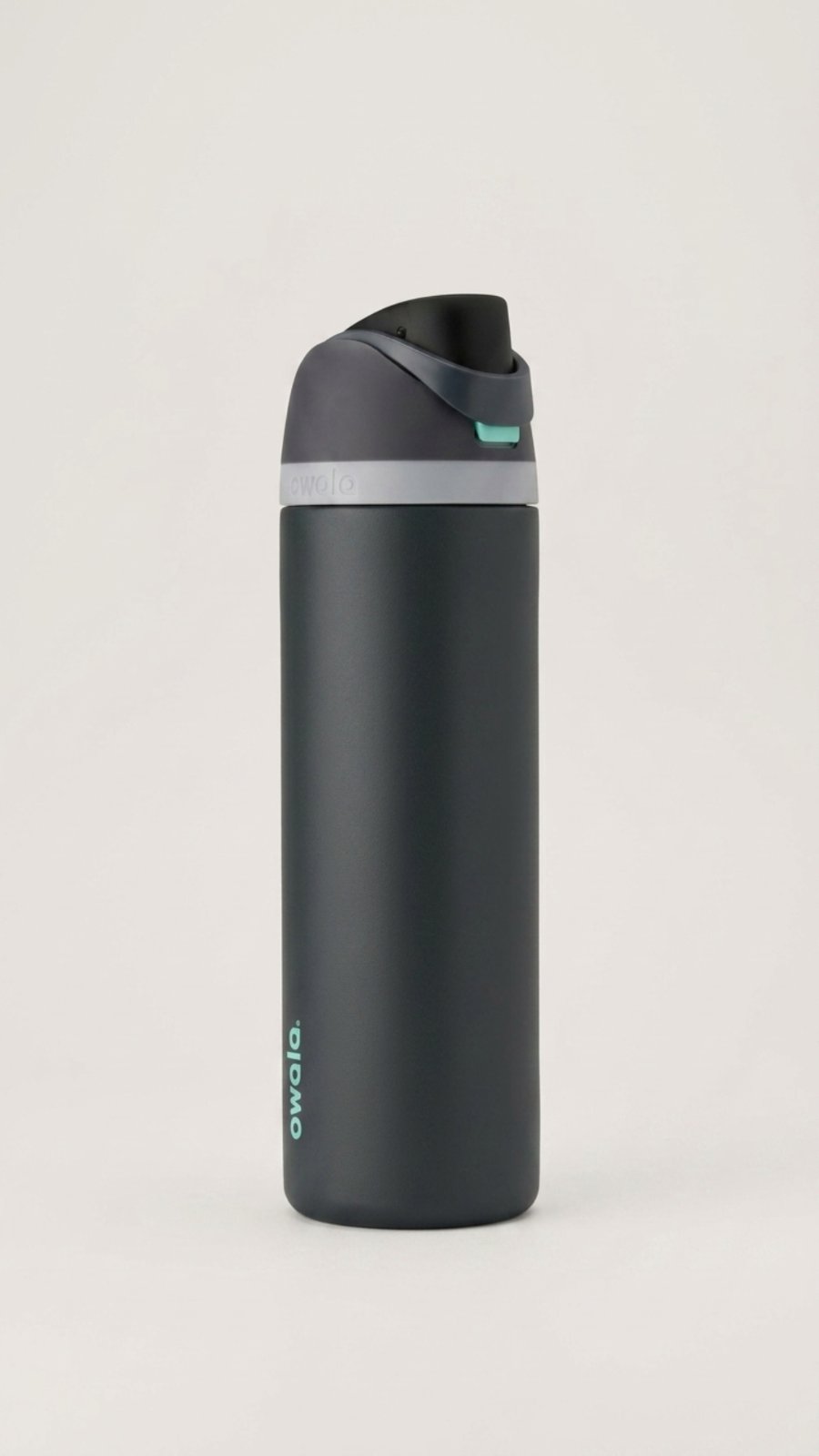 A matte charcoal gray Owala FreeSip water bottle stands upright against a plain light gray background. The bottle has a dark gray lid with a light gray band and a mint green release button.
