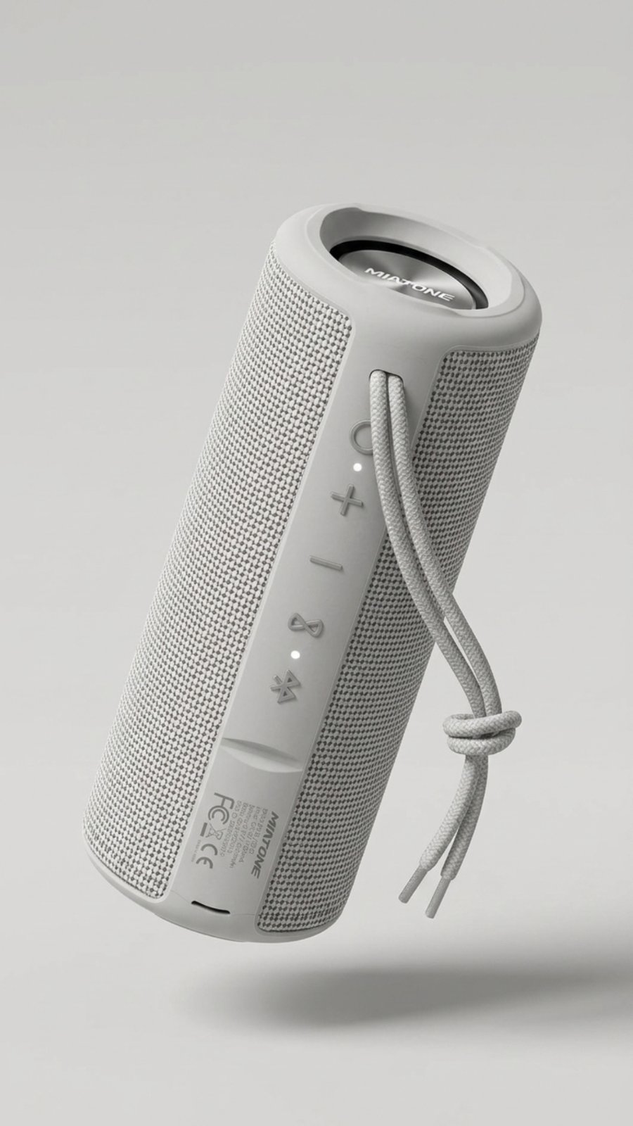 A light gray cylindrical portable speaker with a woven texture body and matte end caps, floating against a light gray background. It has a braided gray cord tied in a knot on one side, and control buttons with small indicator lights on the other side.