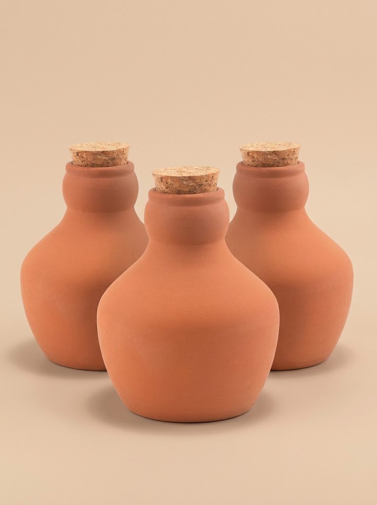 Three terracotta olla watering pots with cork stoppers, arranged in a triangle on a beige background.