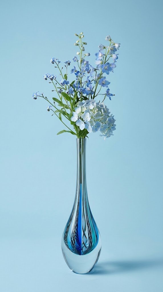 A tall, slender clear glass vase with a teardrop base. The base contains swirling patterns of light blue, dark blue, black, and white. The vase holds light blue and white flowers against a light blue background.