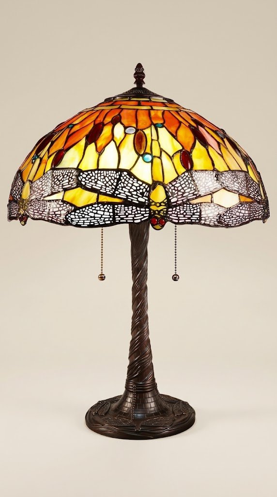 A Tiffany style table lamp with a stained glass shade featuring a dragonfly pattern in amber yellow and red. The lamp sits on a dark surface against a neutral wall.