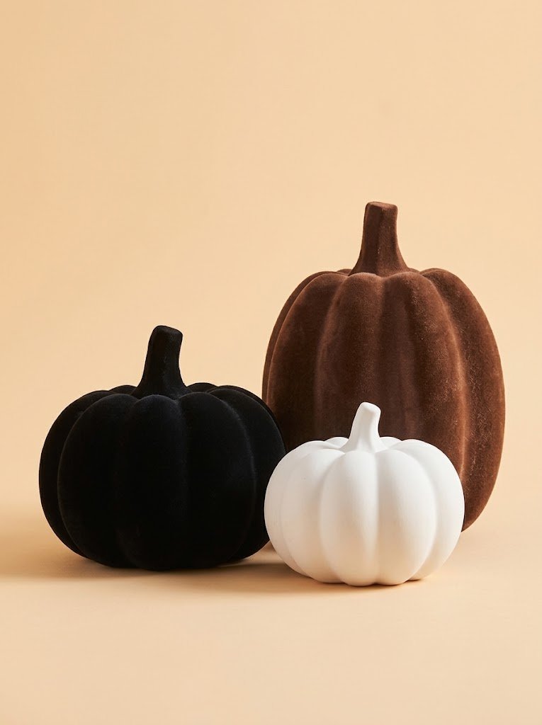 Three flocked pumpkins in white, black, and brown arranged on a wooden surface, each with a rounded shape and small faux stem.