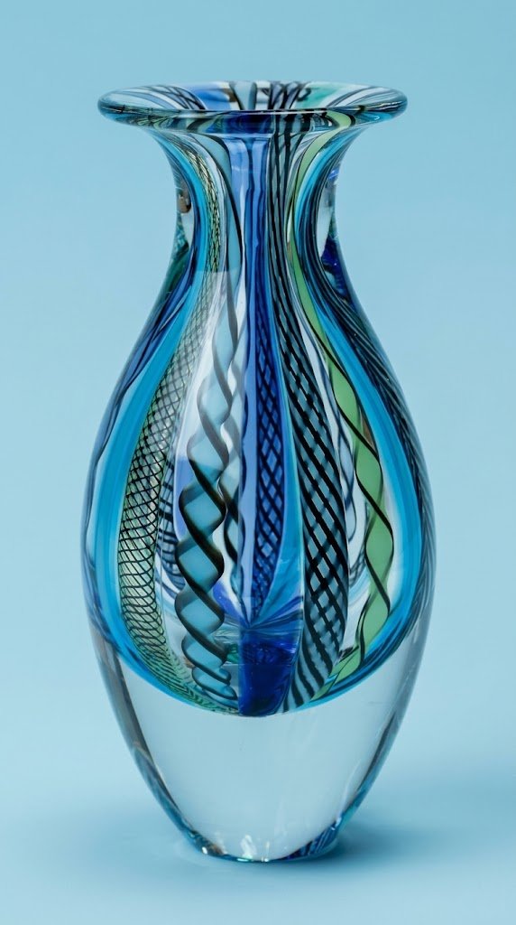 A hand blown teardrop shaped glass vase with vertical ribbons of blue, green, and black mesh canes encased in clear glass on a light blue background.